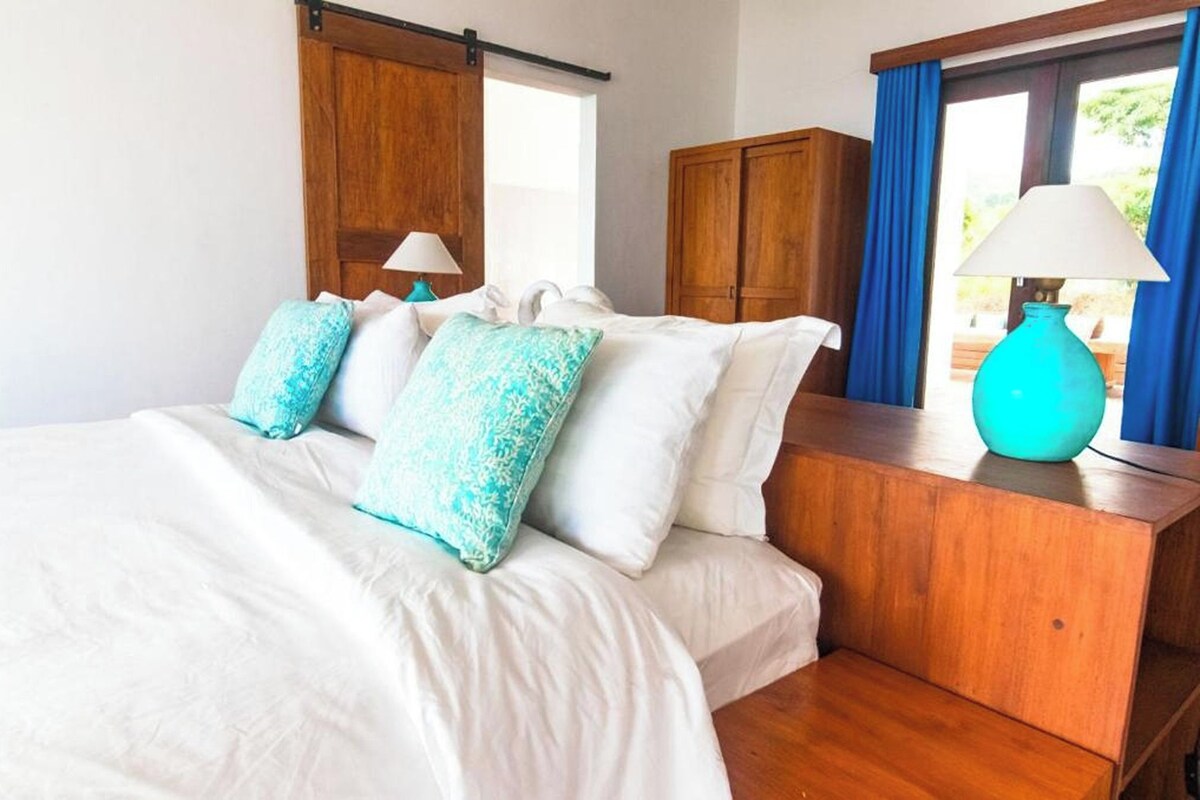 Un dormitorio con cama, cabecera de madera, dos ventanas con cortinas, una mesita de noche con lámpara y vistas al exterior con vegetación.