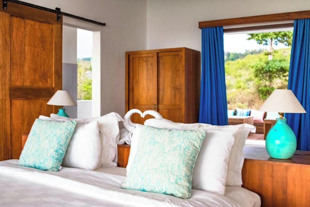 A bedroom with a bed, pillows, a nightstand, a lamp, a wardrobe, and a view of greenery outside.