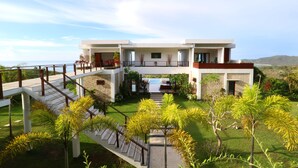 A modern two-story house with a pool, surrounded by lush greenery and a clear view of the ocean.