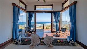 A room with a large balcony, two chairs, a ceiling fan, and a view of the ocean.