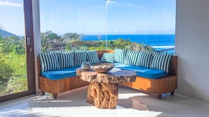A modern room with a large window overlooking a coastal landscape, featuring a wooden bench with blue cushions and a central table.