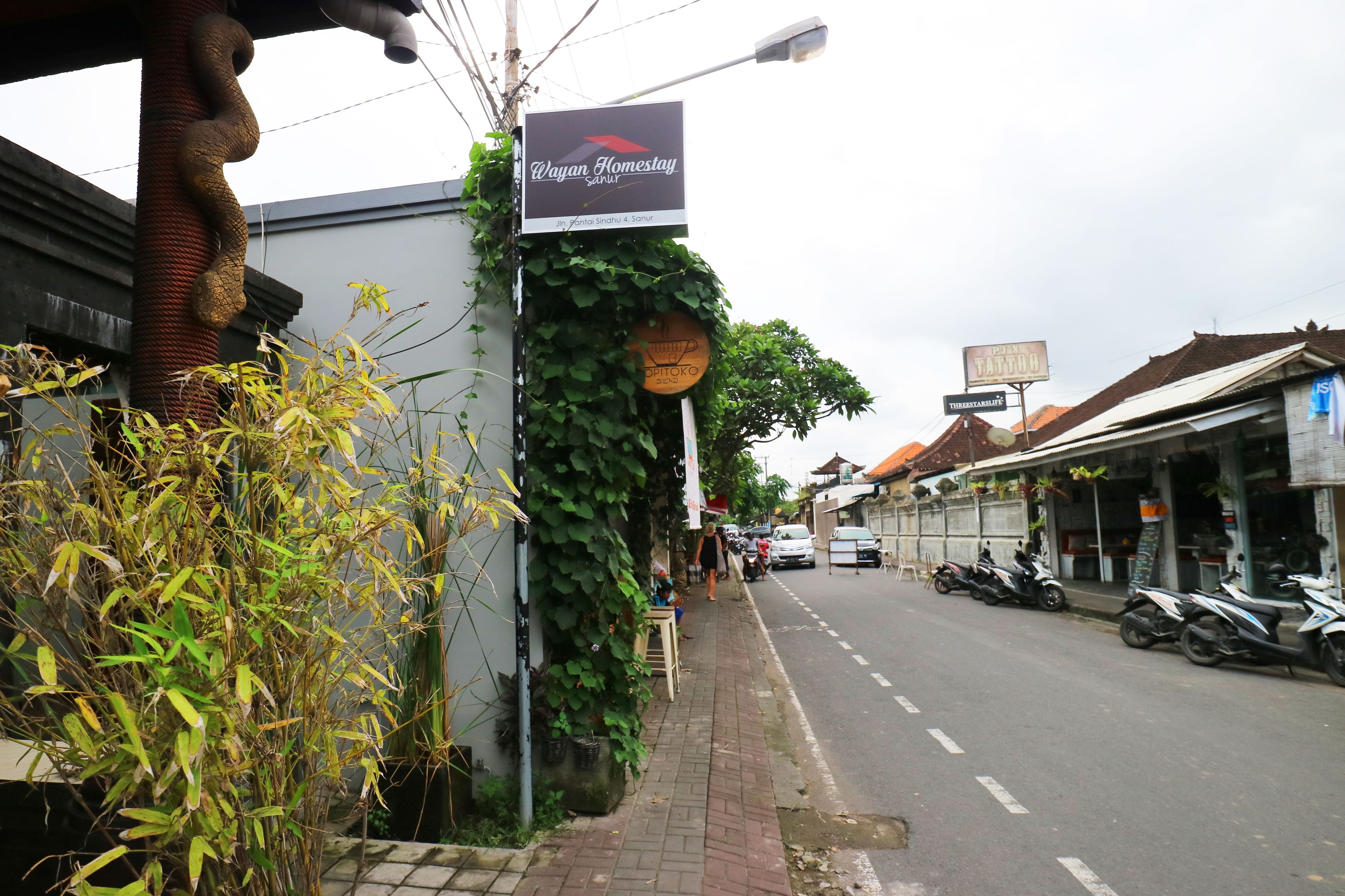A street with a sign for Wayan Homestay, a building with a sign for a tattoo shop, and parked motorcycles.