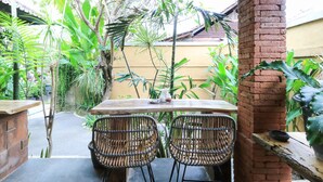 Une terrasse extérieure avec une table en bois et des chaises en osier, entourée d’une végétation luxuriante et d’un pilier en briques.