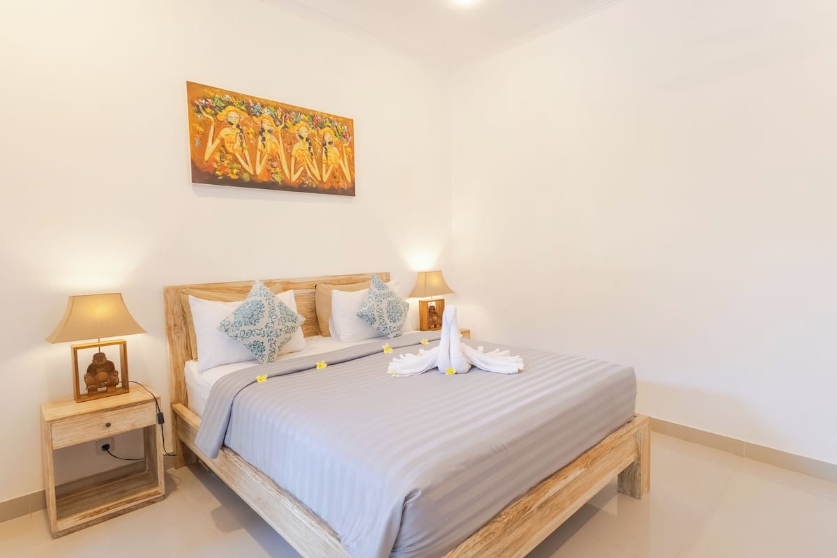 A neatly made bed with a grey bedspread, two bedside lamps, and a wooden nightstand with a small Buddha statue.