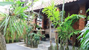 Un bâtiment en briques surmonté d’un toit en tuiles, entouré de diverses plantes tropicales et doté d’un espace d’entrée pavé.