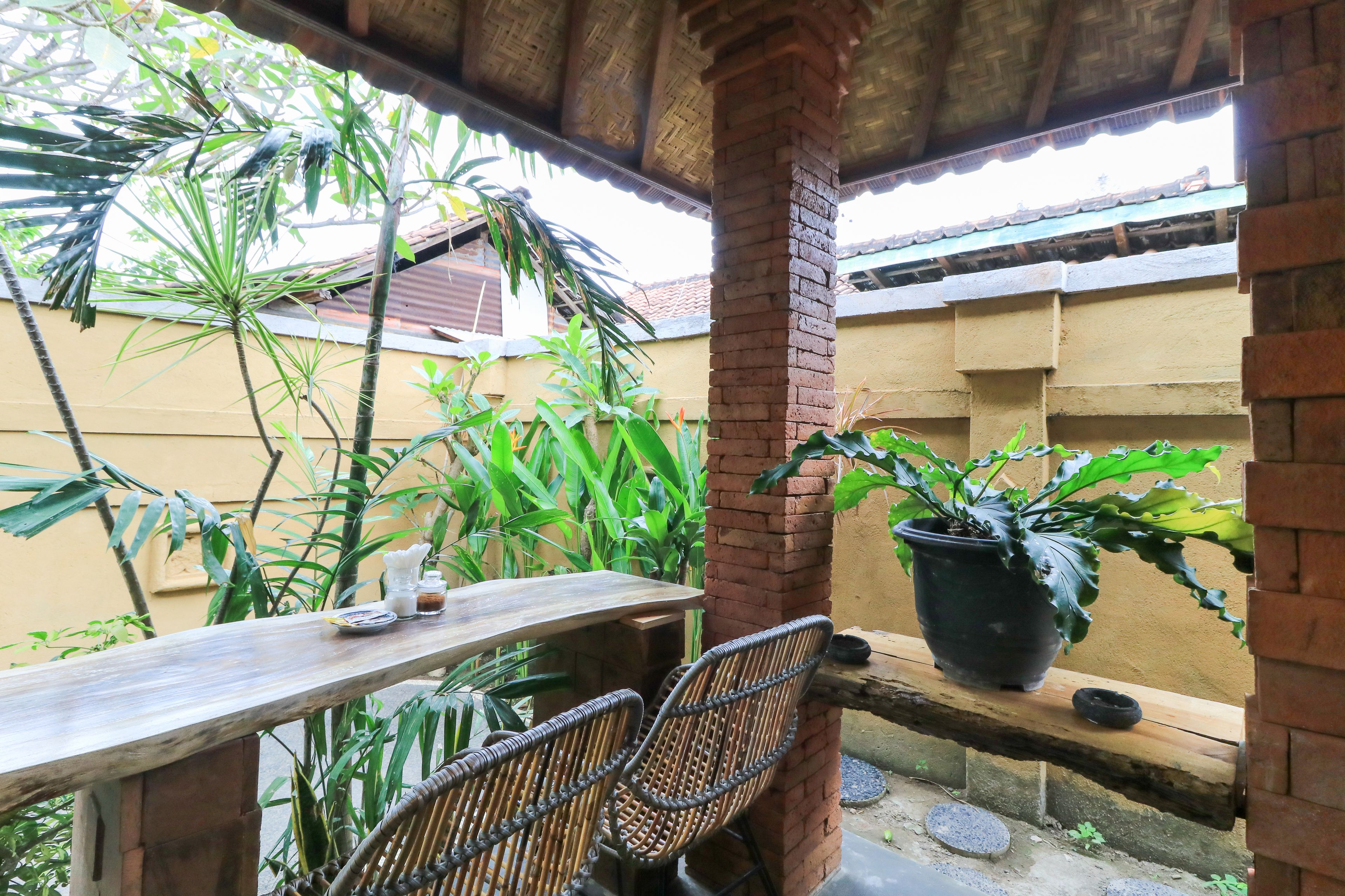 A covered outdoor seating area with a wooden table and wicker chairs, surrounded by potted plants and a brick wall.