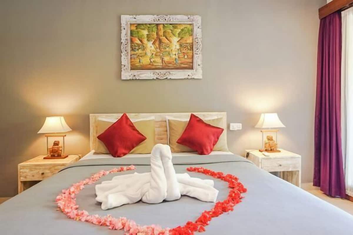 A neatly made bed with a heart-shaped arrangement of towels and flowers, two red pillows, and a painting on the wall.