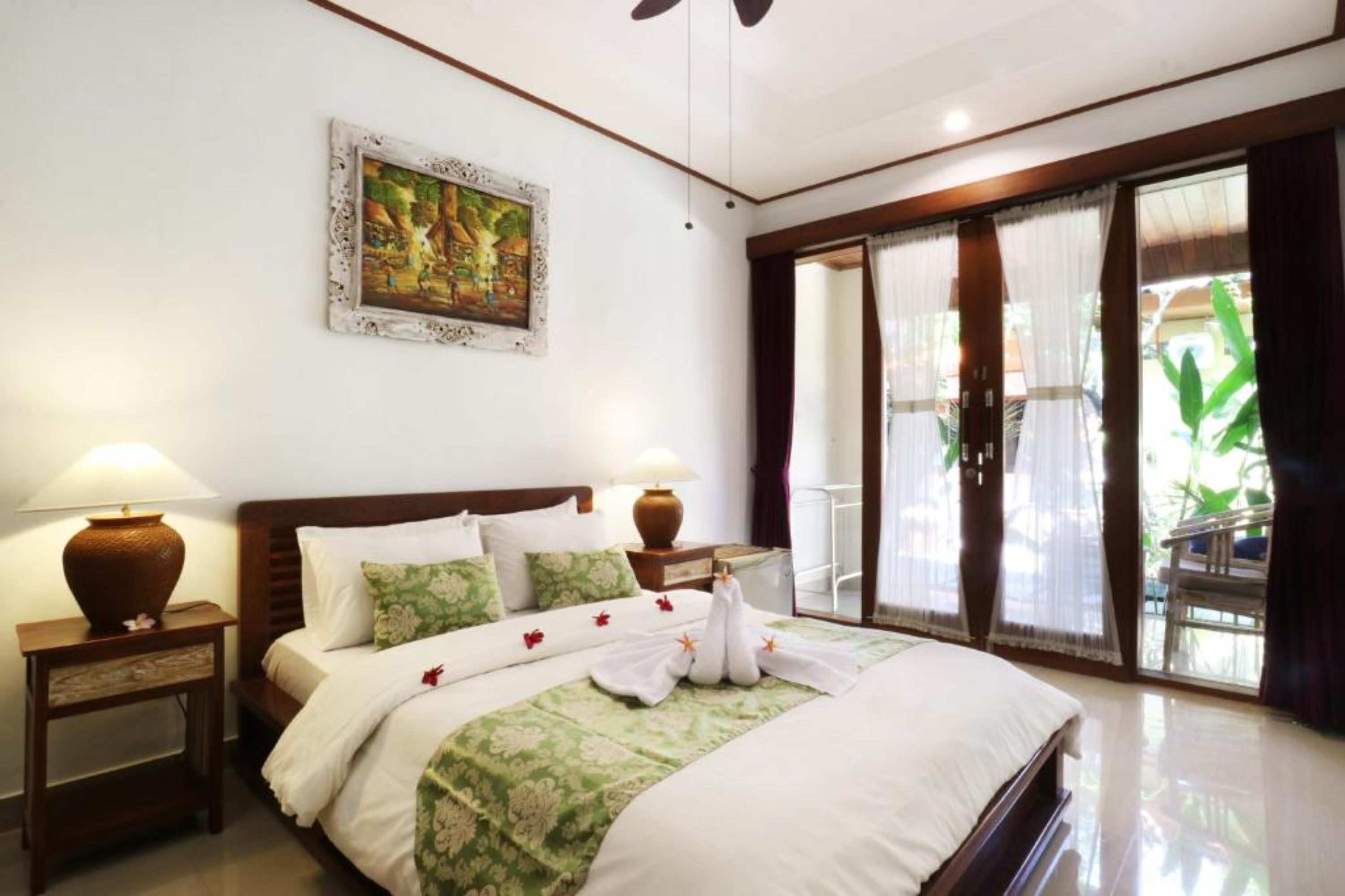 A hotel room with a large bed, two bedside tables, a painting on the wall, and a view of the outdoors through a sliding door.