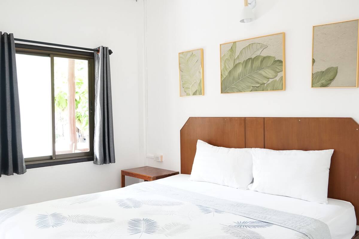 Ein Schlafzimmer mit Holz-Kopfseitenverkleidung, einem Bett mit weißer Bettwäsche, einem Nachttisch und einem Fenster mit Vorhängen. An der Wand hängen drei gerahmte Bilder mit Blättern.