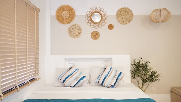 A bedroom with a bed, a window with blinds, and wall decor including woven wall hangings and a mirror. - Yoghurt Home (Ko Pha-ngan)