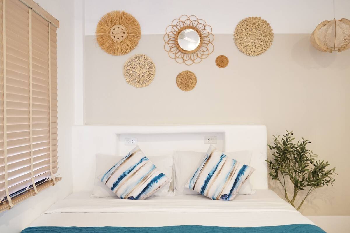 Ein Schlafzimmer mit einem Bett, einem Fenster mit Jalousien und Wanddekoration, darunter Wandbehänge und ein Spiegel.