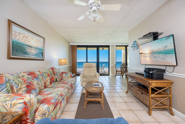 A living room with a sofa, a chair, a television, and a view of the ocean through large windows.