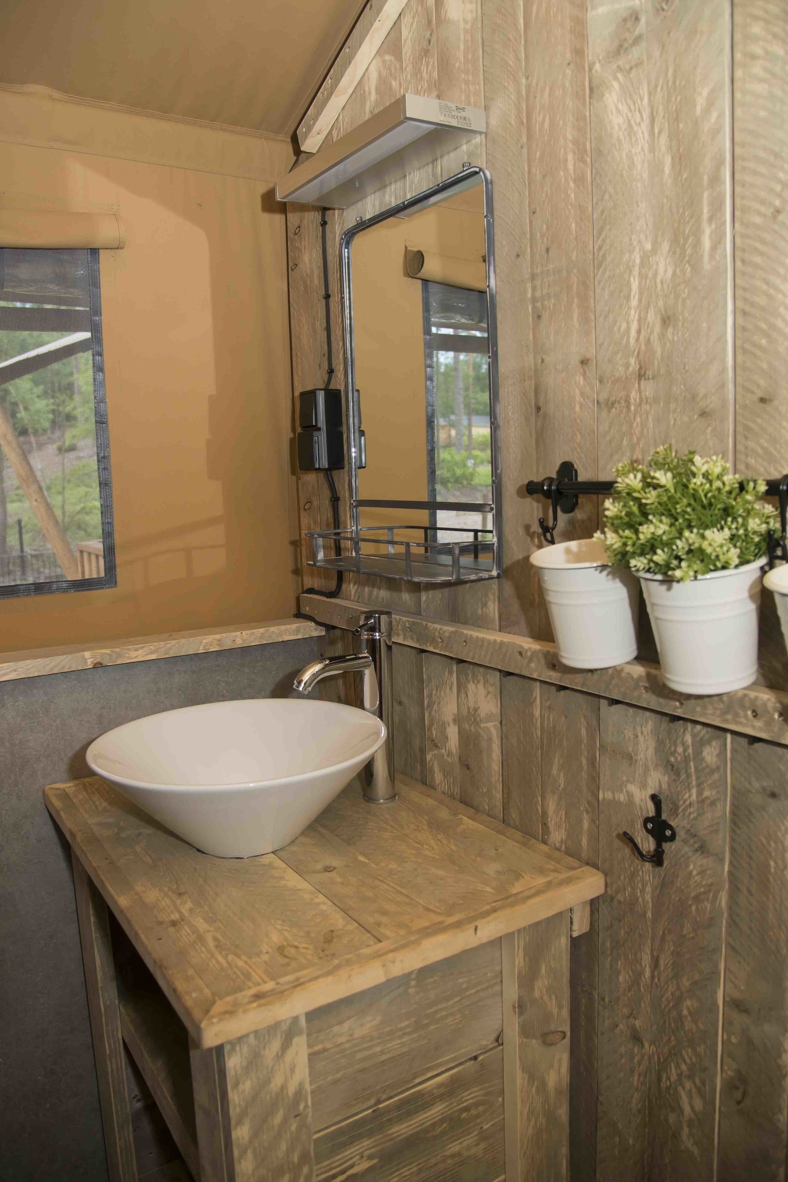 A rustic bathroom with a wooden vanity, a white vessel sink, a mirror, and two potted plants.