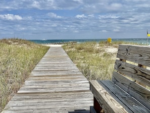 En træbro fører hen til en strand, og der er en bænk til højre.