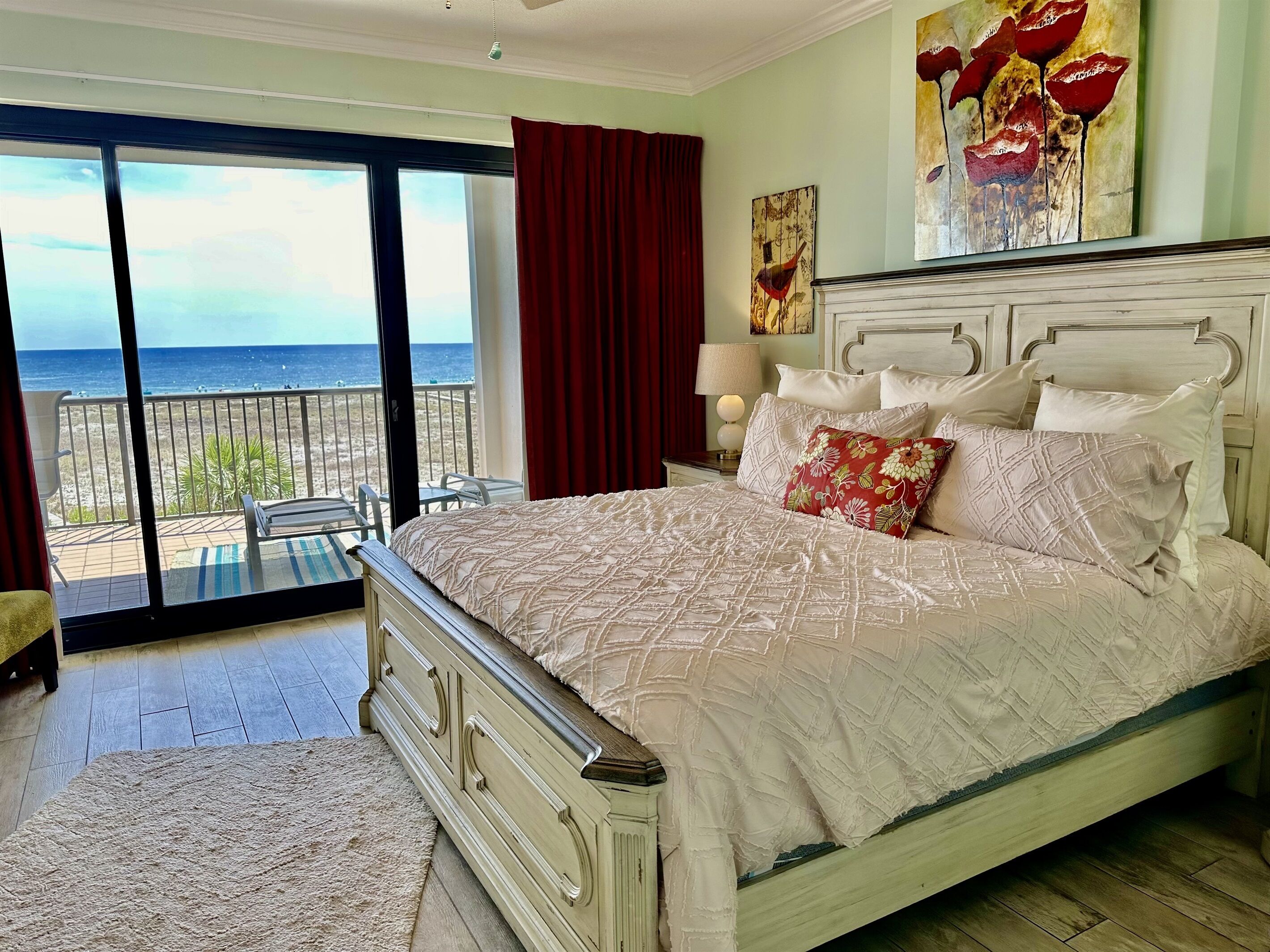 A bedroom with a large bed, a nightstand, a lamp, and a painting. There is a balcony with a view of the ocean.