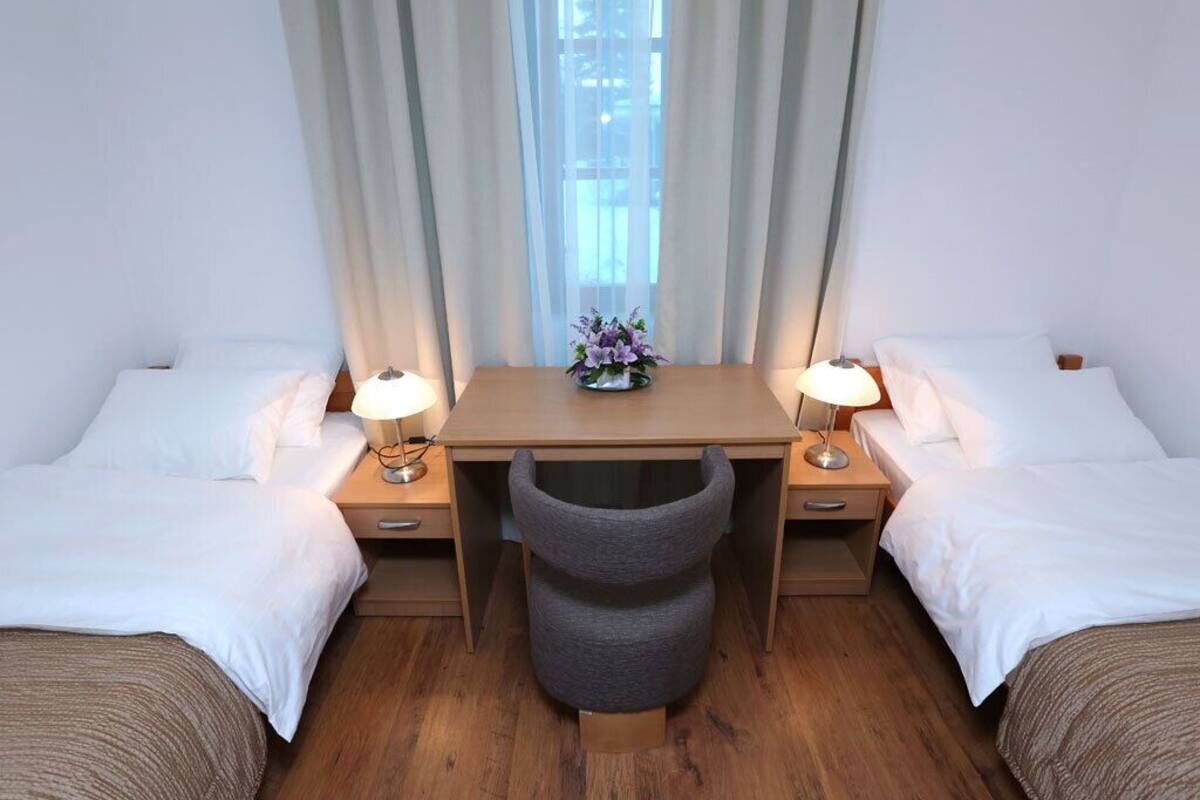 A hotel room with two single beds, a wooden desk with a chair, a small table with a lamp, and a window with curtains.