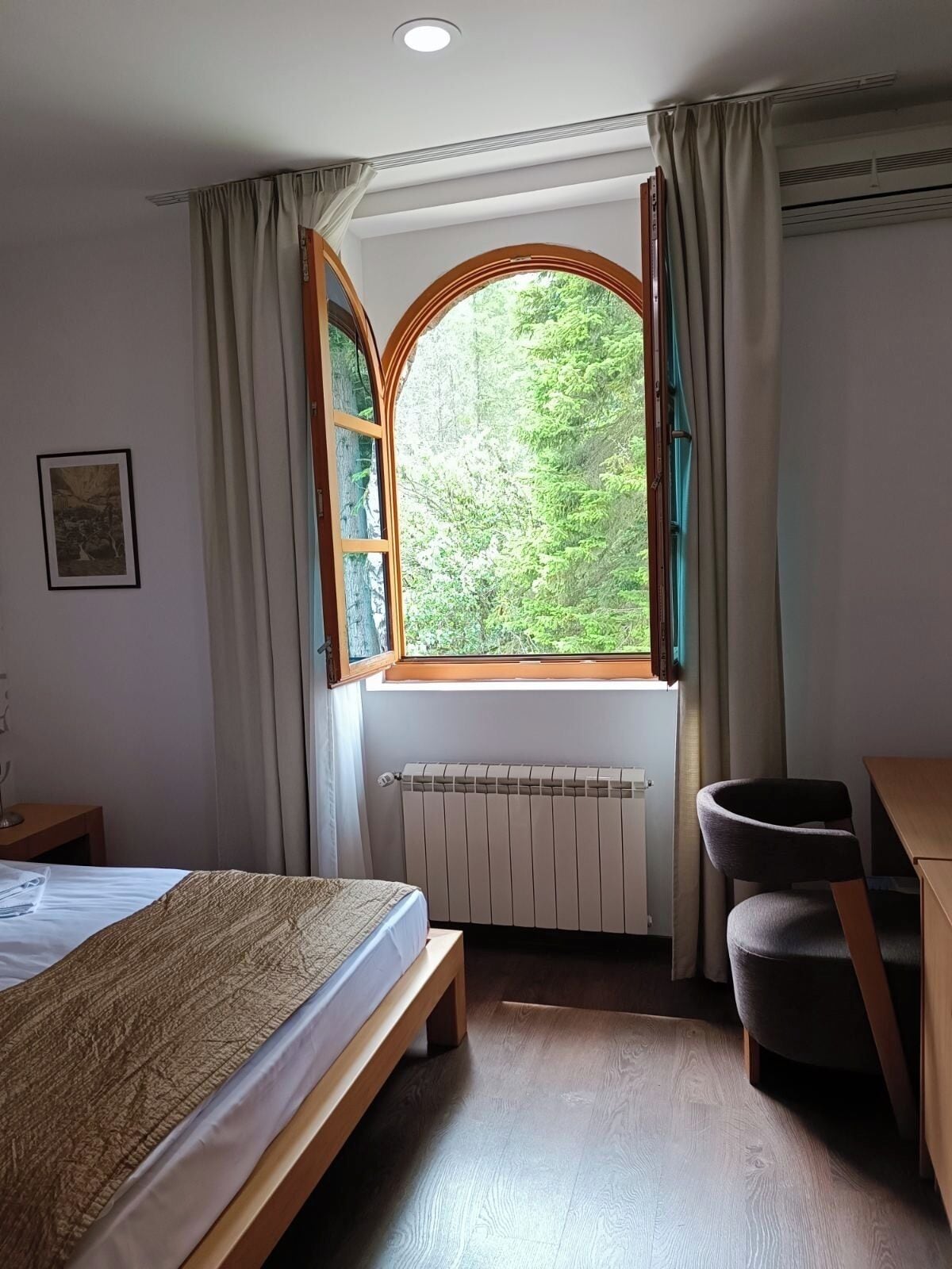 A bedroom with a bed, a desk, a chair, a radiator, and a window with curtains.