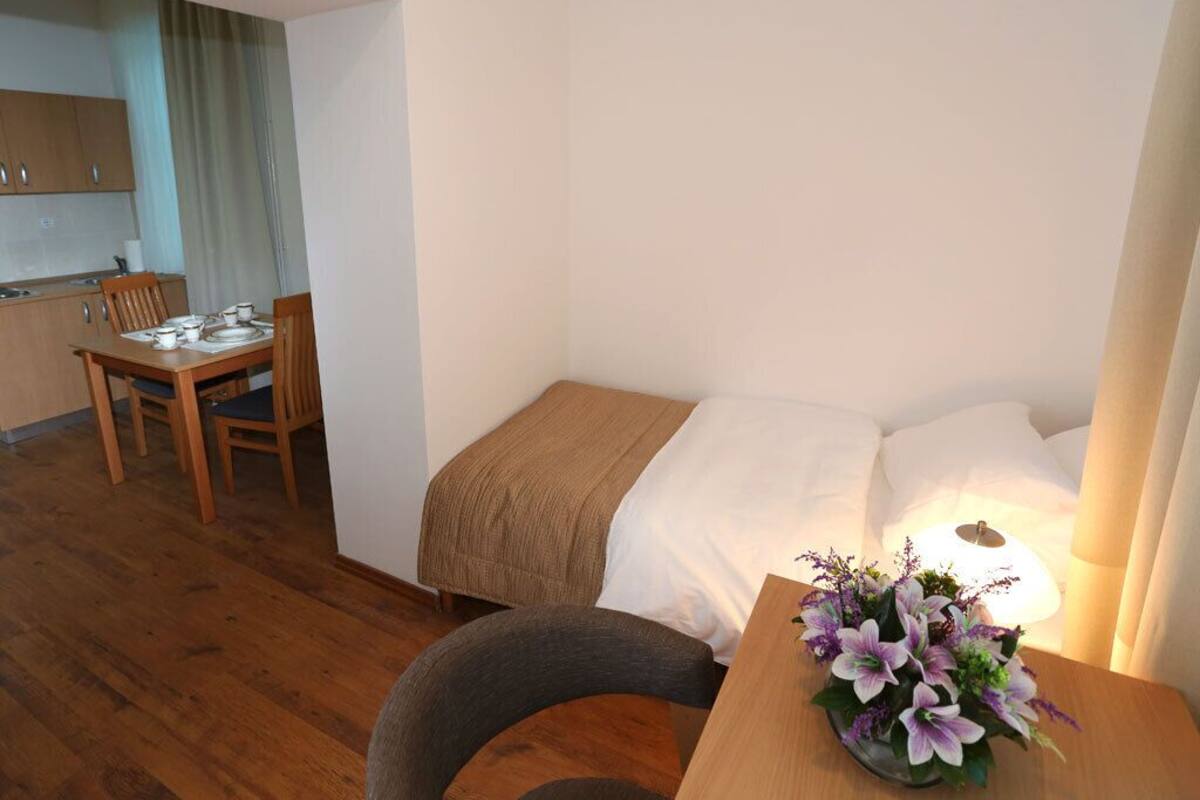 A compact hotel room with a single bed, a bedside table with a lamp, a small desk, and a chair. There is a kitchenette with a table and chairs in the background.