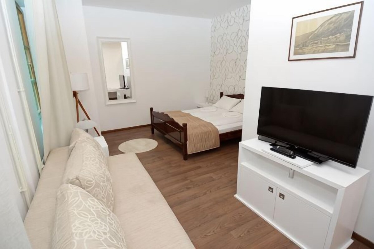 Un dormitorio con cama, televisor, sofá, espejo y ventana.