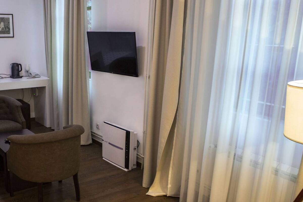 A hotel room with a bed, a desk with a computer, a chair, a TV mounted on the wall, and a window with sheer curtains.