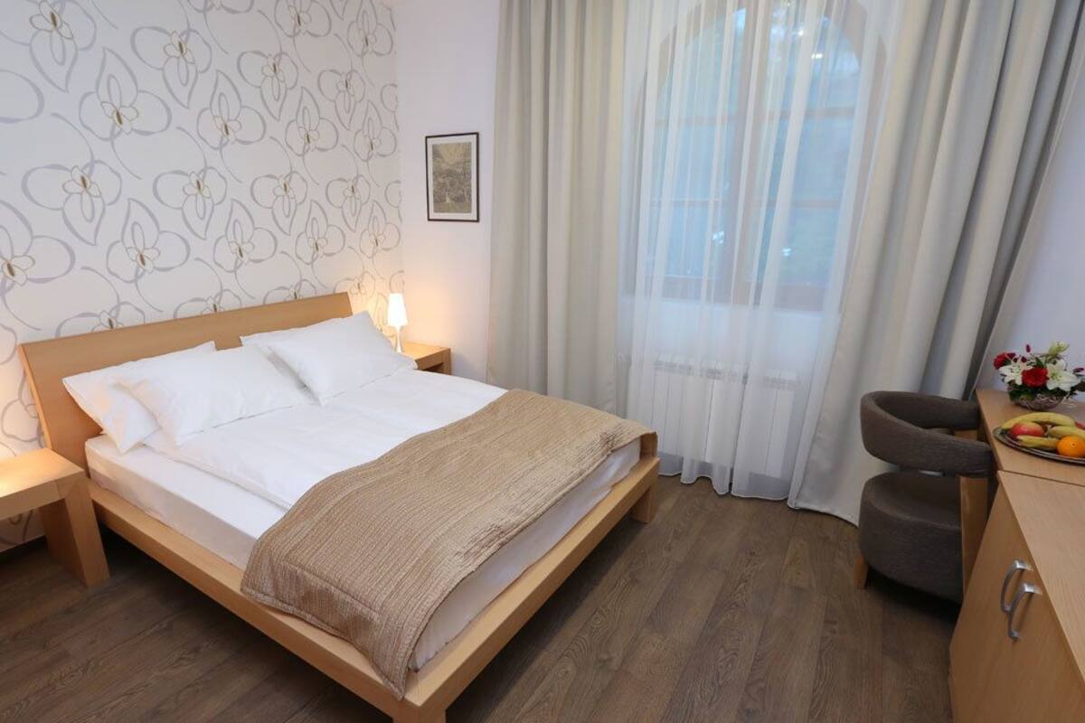 A bedroom with a wooden bed, a nightstand, a chair, a desk with a fruit bowl, and a window with sheer curtains.