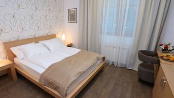 Ein Schlafzimmer mit einem hölzernen Bett, einem Nachttisch, einem Sessel, einem kleinen Tisch mit Früchten und einem Fenster mit Vorhängen.