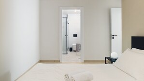 A minimalist hotel room with a large bed, a bedside table with a lamp, and a door leading to a bathroom. - Elegant Duplex with XL Patio by FeelHome (Tel Aviv)