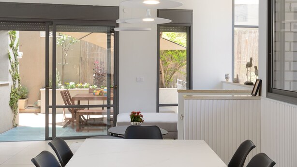 A modern dining area with a white table and black chairs, large windows, and a view of the outdoor patio. - Elegant Duplex with XL Patio by FeelHome (Tel Aviv)