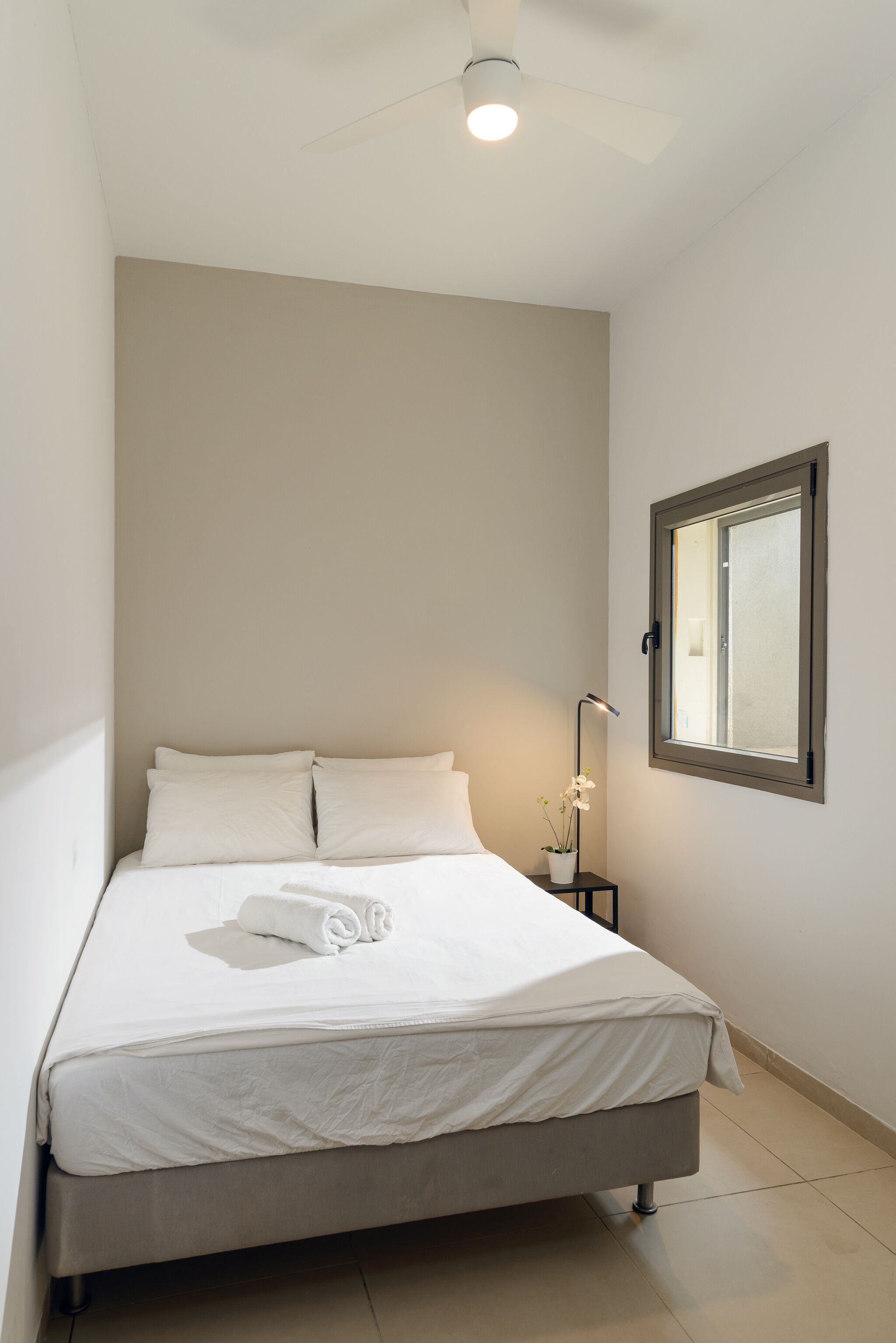 A neatly made bed with white linens and a rolled towel, a bedside table with a lamp, and a window with blinds.