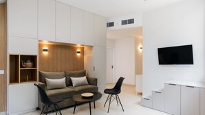 A modern living room with a sofa, two chairs, a coffee table, built-in shelving, a flat-screen TV, and a staircase leading to another level. - Elegant Duplex with XL Patio by FeelHome (Tel Aviv)