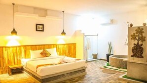 Una habitación de hotel con cabecera de madera, cama con ropa de cama blanca, dos lámparas colgantes, un baño pequeño con ducha y una planta decorativa.