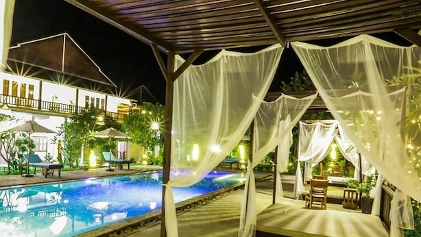 Zona de piscina nocturna con área de estar cubierta y un edificio blanco al fondo.