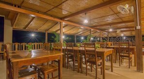 Un restaurante con mesas y sillas de madera, techo de paja y vistas al cielo nocturno.