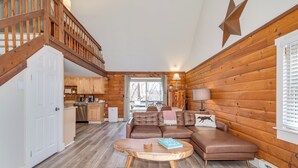 A cozy cabin interior with a wooden staircase, a living area with a brown leather sofa, a round coffee table, and a kitchen in the background. - Secluded Pet-Friendly High-Tech Log Cabin in the Woods (Berkeley Springs)