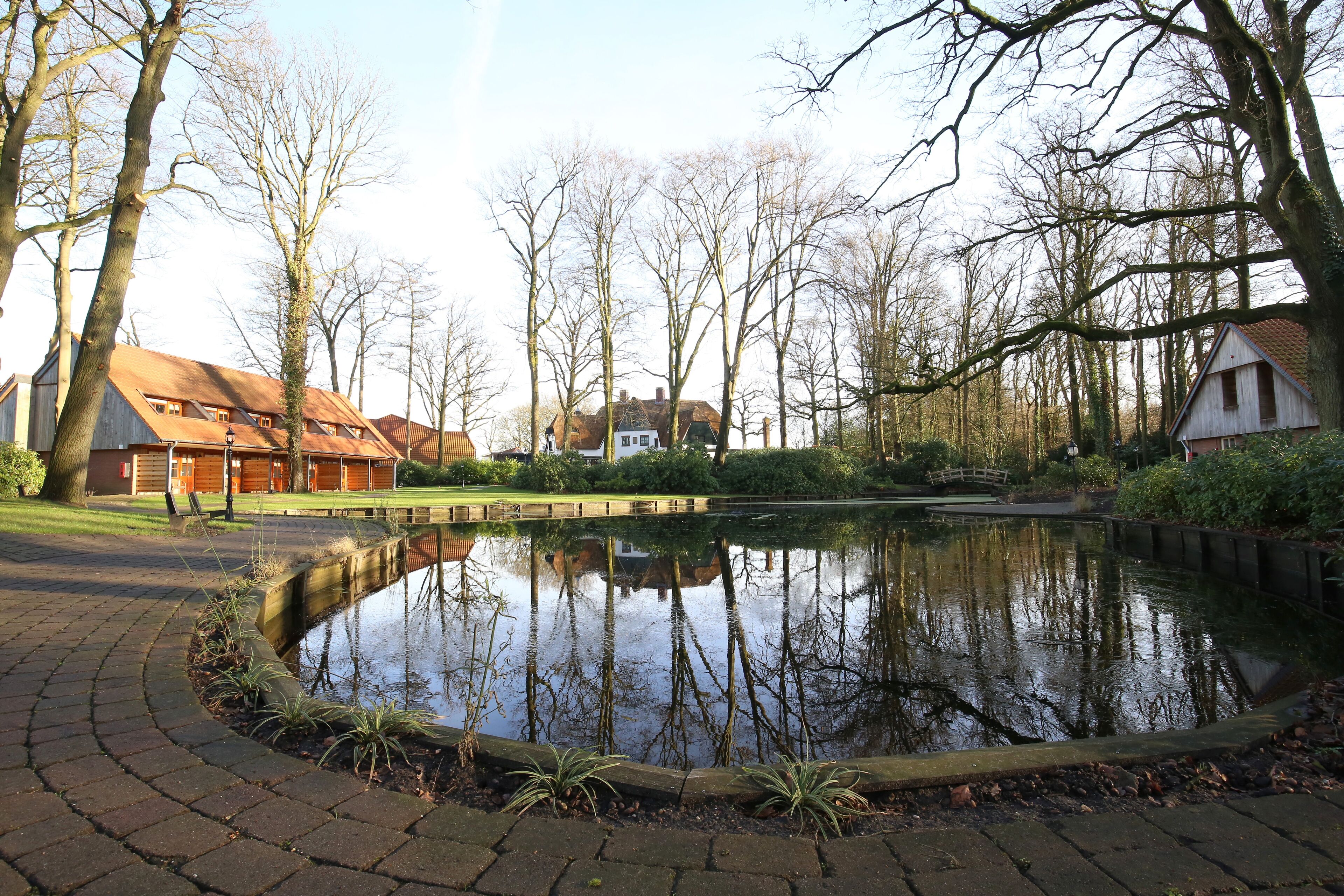 A pond with a wooden bridge, surrounded by trees and a building with a red roof.