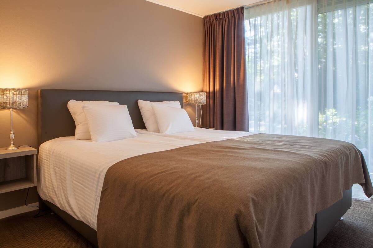 Una cama bien hecha con sábanas blancas y un edredón marrón, con dos mesitas de noche a cada lado, cada una con una lámpara, en una habitación con un ventanal y cortinas de encaje.