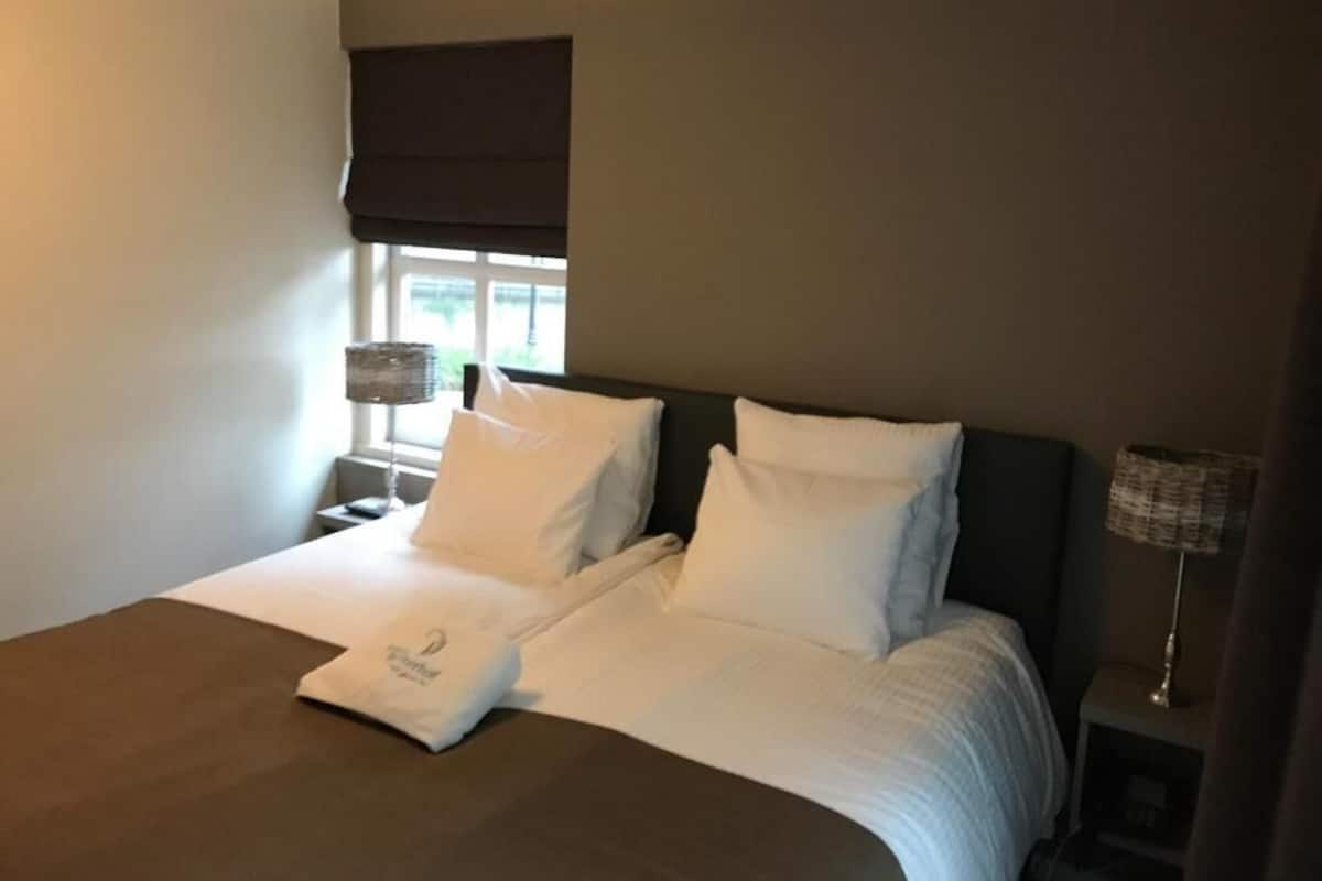 Una cama bien hecha con sábanas y almohadones blancos, una mesita de noche con lámpara, y una ventana con persiana oscura.
