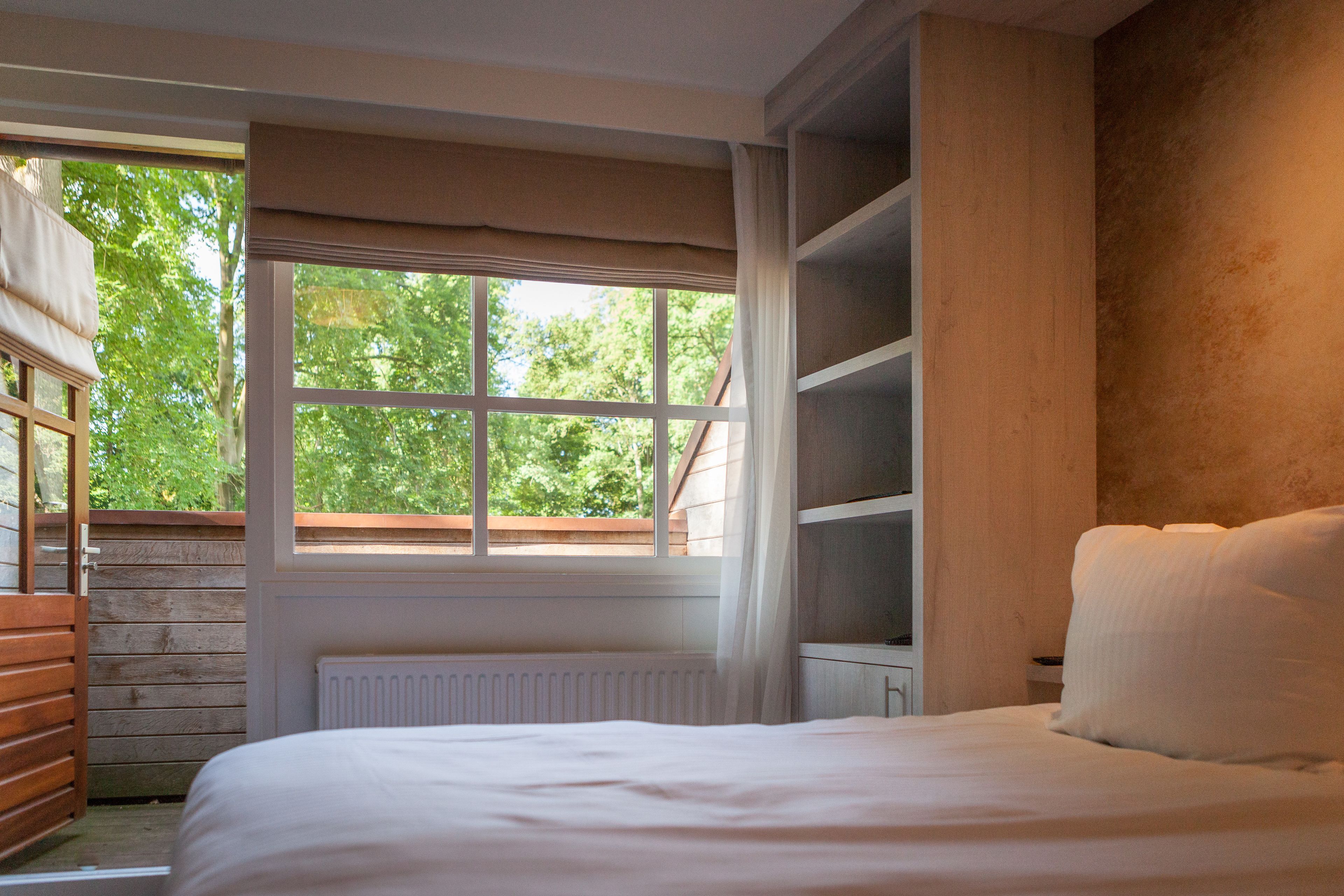 A bedroom with a large window, wooden slatted walls, a white radiator, and a bed with white bedding.