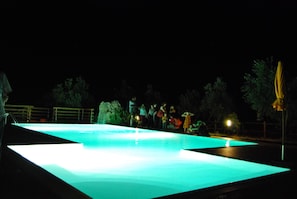 En pool som används för simning på natten, med en upplyst kant, omgiven av ett staket och träd.