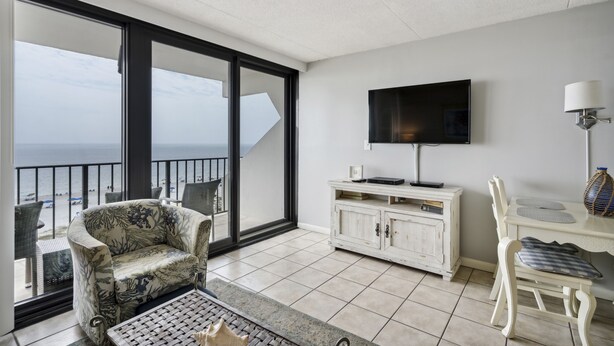 A balcony with a view of the sea, a sofa, a TV, and a dining table with chairs. - Island Winds East 505 by Liquid Life (Gulf Shores)