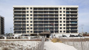 A multi-story hotel with a central entrance, surrounded by a sandy beach and a wooden fence. - Island Winds East 505 by Liquid Life (Gulf Shores)