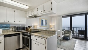 A modern kitchen with white cabinetry, a granite countertop, and stainless steel appliances. There is a dining area with a view of the ocean through large windows. - Island Winds East 505 by Liquid Life (Gulf Shores)