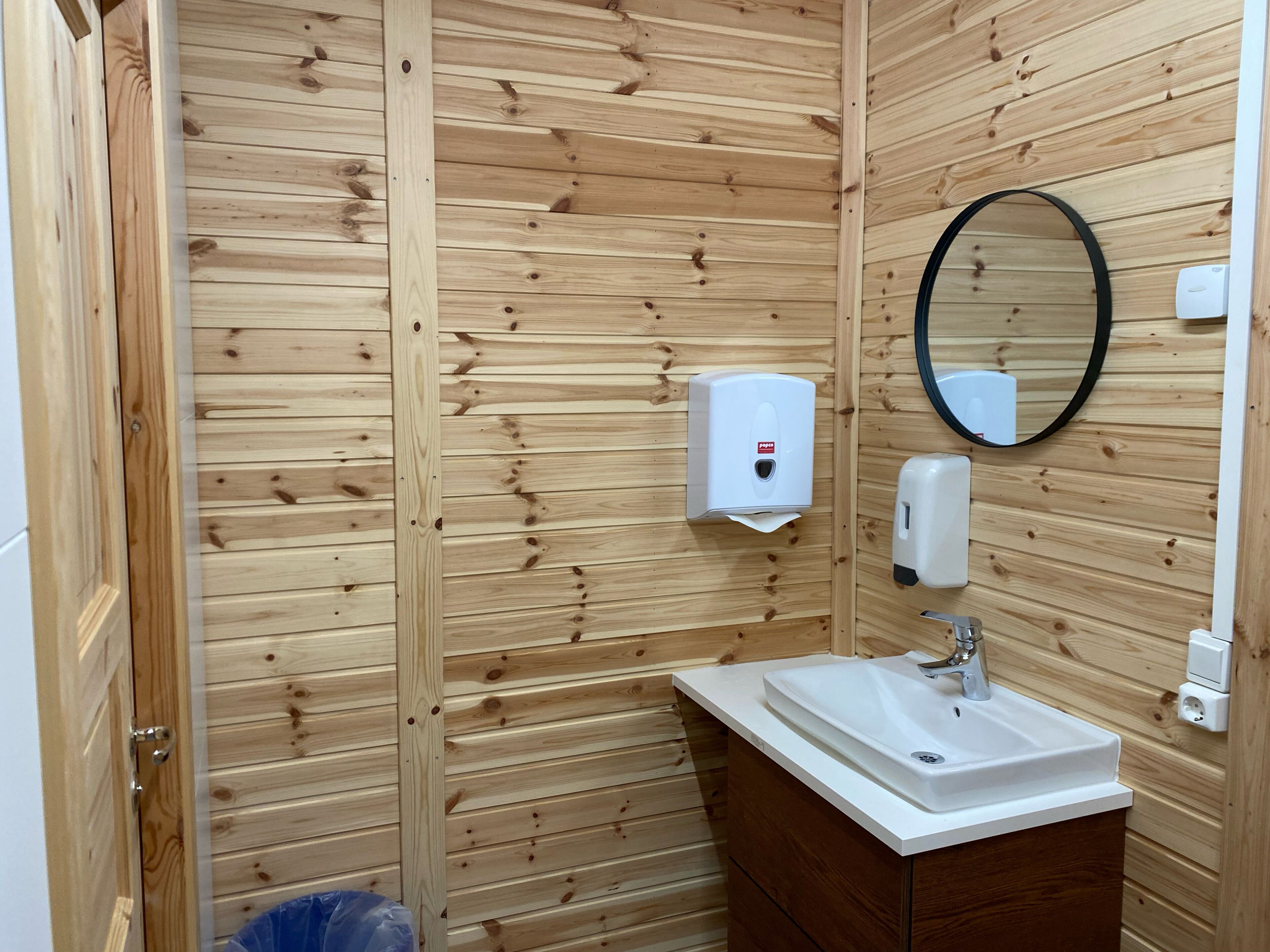 A wooden-paneled bathroom with a sink, mirror, soap dispenser, and toilet paper holder.