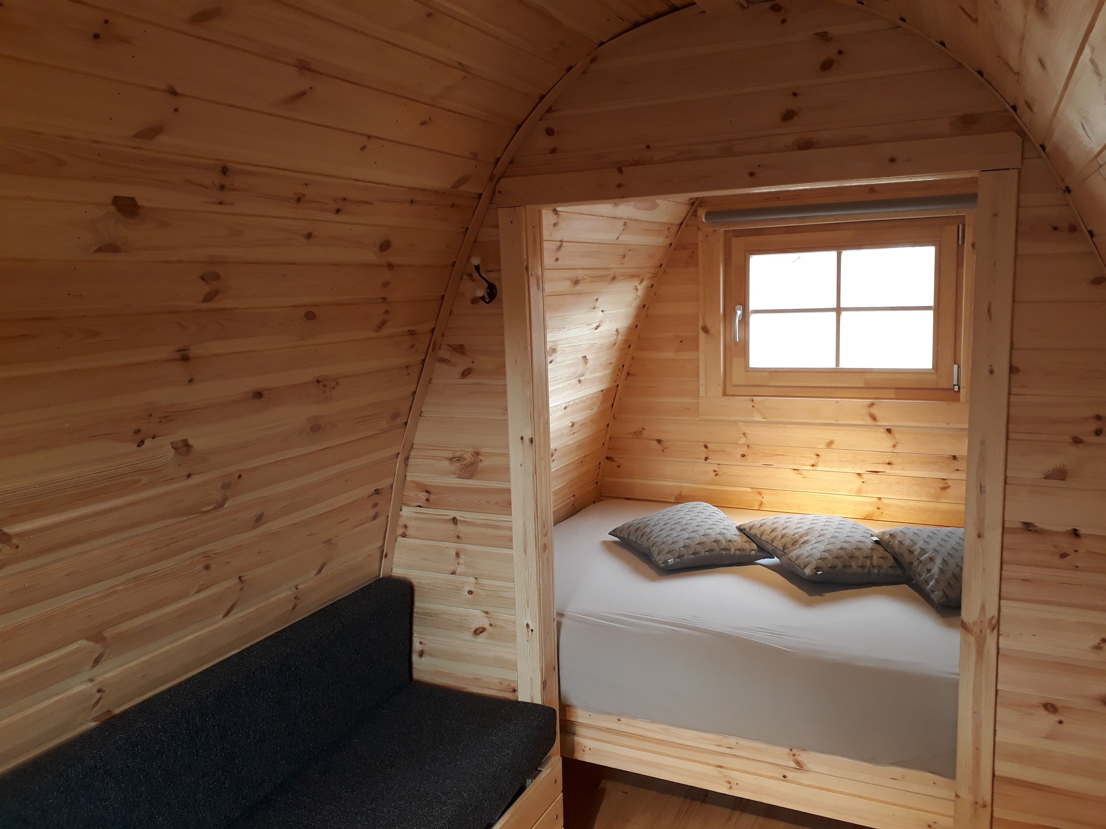 A small, cozy room with wooden walls and a sloped ceiling. There is a bed with pillows and a bench against the wall. A window allows natural light to enter.