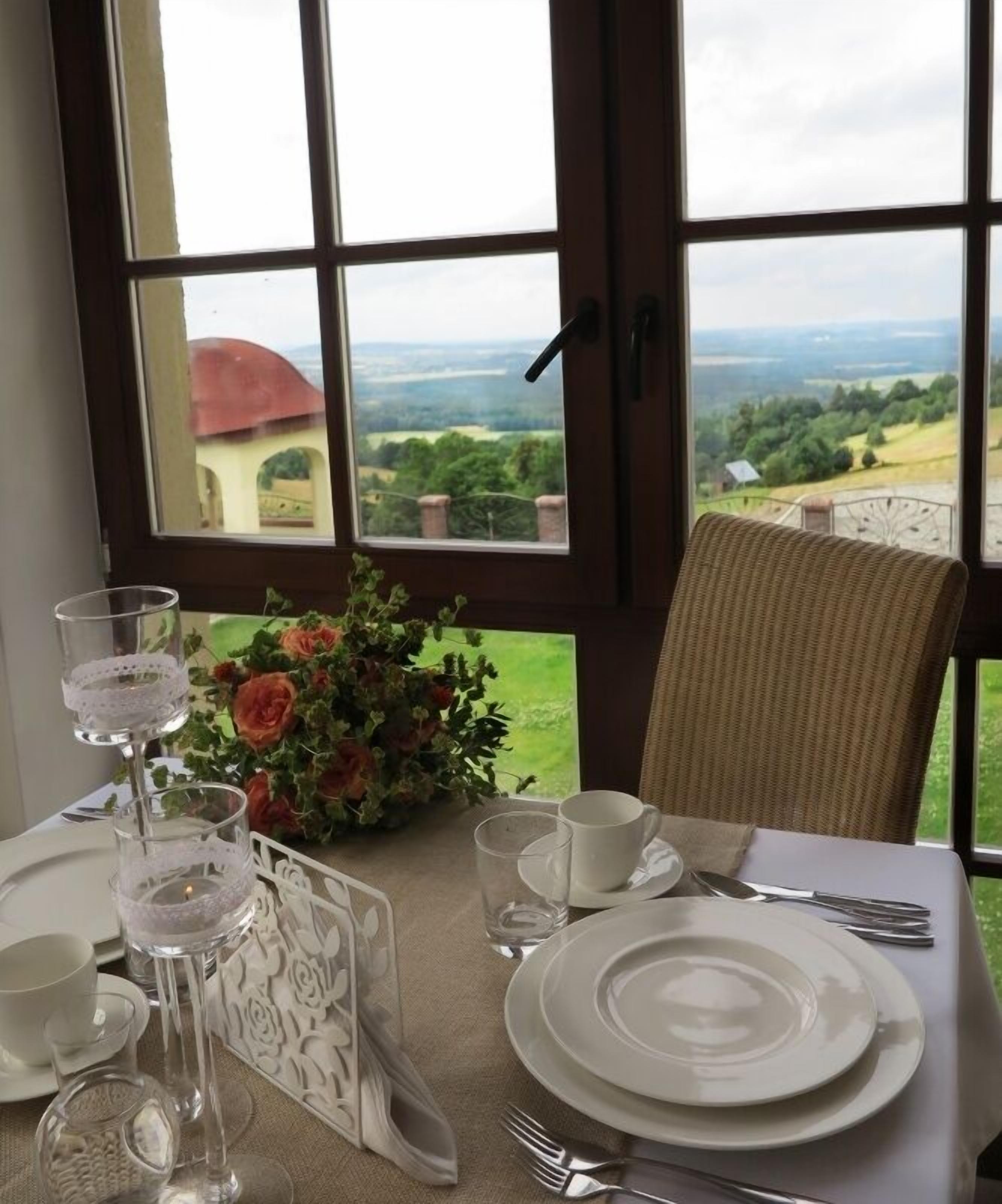 A dining table set for two with a view of a countryside landscape through large windows.