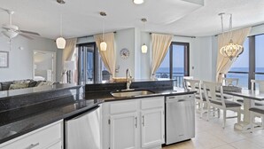 Een moderne keuken met een zwart aanrecht, witte kasten en een ingebouwde vaatwasser. Er is een eethoek met witte stoelen en een tafel, en grote ramen met uitzicht op de oceaan.