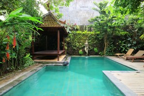 A pool area with a wooden deck, lounge chairs, and a small pavilion surrounded by lush greenery.