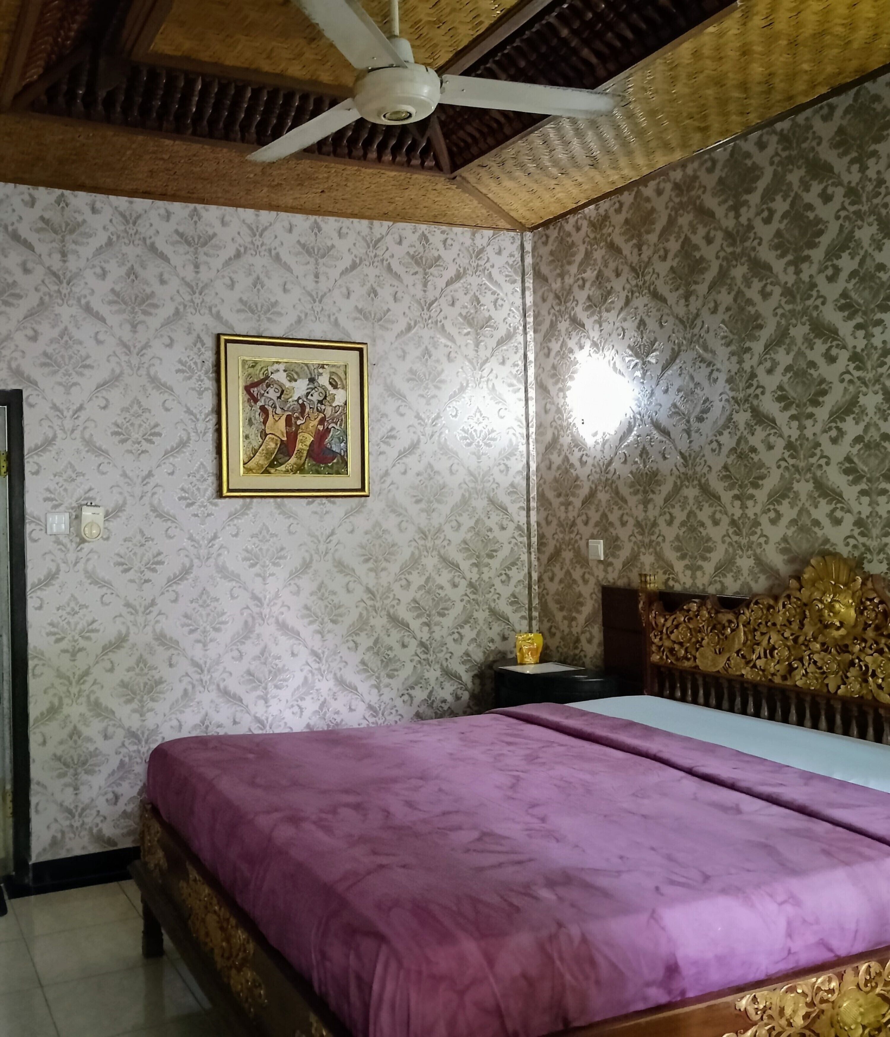 A room with a bed, a ceiling fan, a framed picture on the wall, and patterned wallpaper.
