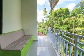 A balcony with a cushioned bench, white railing, and a view of lush greenery and a building in the background.