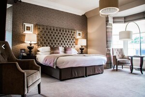 Quarto de hotel com uma cama grande, duas luminárias de cabeceira, uma cabeceira estofada, uma cadeira e uma mesa.